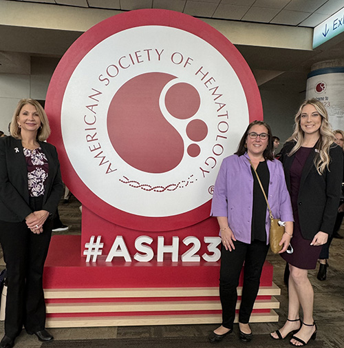 Caroline, Jenn, and Melissa standing next to the ASH 2023 conference sign